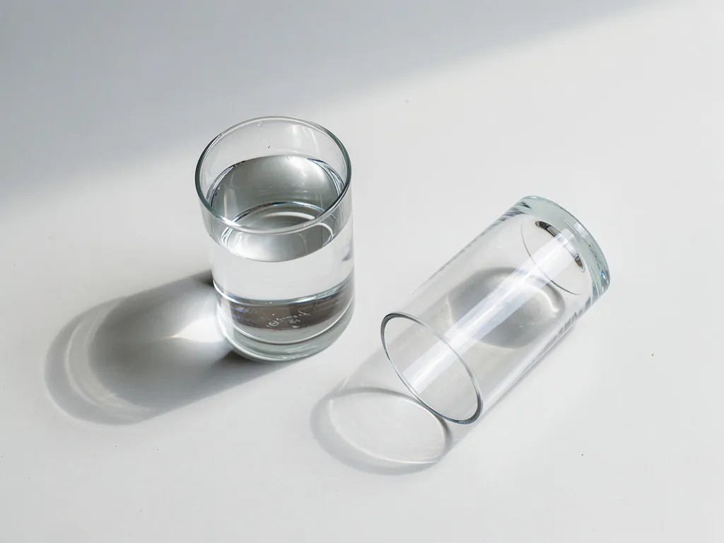 Twee glazen water op witte ondergrond, één rechtop gevuld en één omgevallen leeg, minimalistische compositie met zachte schaduwen